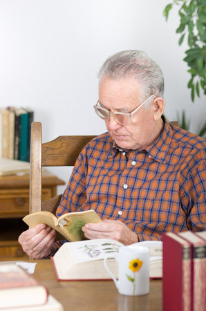 Old man with reading glasses reading book in dining roomの写真素材