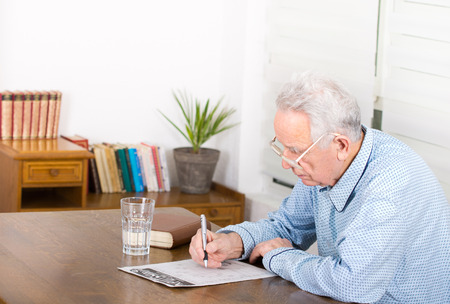 Senior man in pajamas with reading glasses sitting at table and solving crosswordsの写真素材