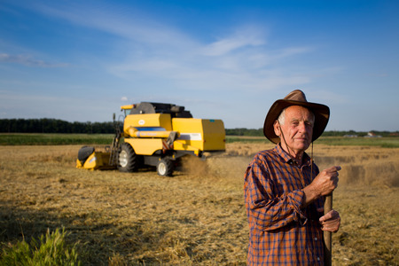 Smiling old peasant standing on field with combine in backgroundの写真素材
