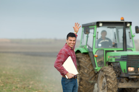 Enthusiastic young farmer waving with hand on farmland with tractor in backgroundの写真素材