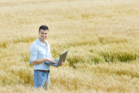Businessman with laptop standing in ripe wheat fieldの写真素材