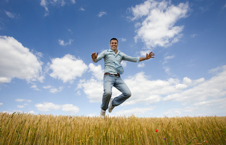 Young attractive man jumping over yellow wheat field blue sky with white clouds in backgroundの写真素材