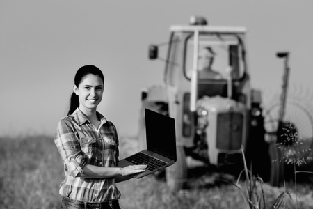 Beautiful young woman with laptop standing in the field with tractor in background, black and white imageの写真素材