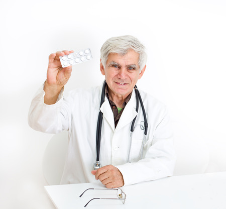 Senior doctor showing blister of pills in raised arm at white tableの写真素材