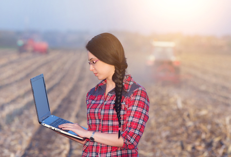 Young woman agronomist standing on field with laptop. Tractors working in backgroundの写真素材