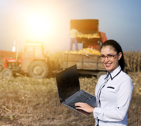 Young woman with laptop standing on corn field. Tractor and harvester working in backgroundの写真素材