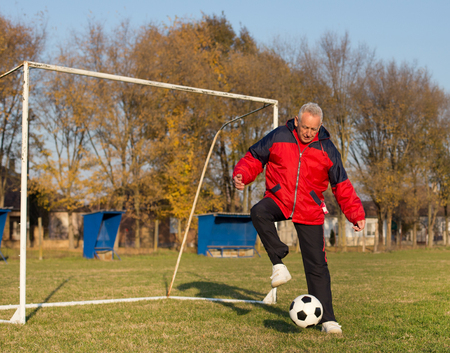 Old man in seventies kicking a soccer ball on playground with goal behind himの写真素材