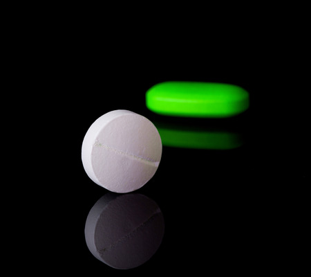 White round tablet with one green pill in background. Black background with reflectionの写真素材