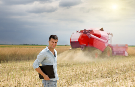 Young attractive businessman standing in the field, combine harvester in backgroundの写真素材
