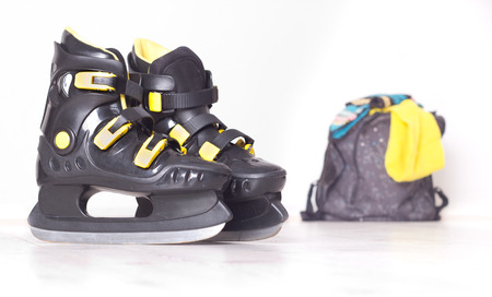 Close up of black plastic ice skates with yellow details and backpack with colorful cap and scarf in backgroundの写真素材