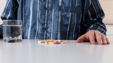 Man in pajamas sitting at white table  with pile of pills and glass of water in front of himの写真素材