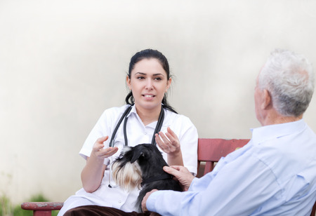 Young pretty nurse explaining something to senior man with dog on the bench in courtyardの写真素材