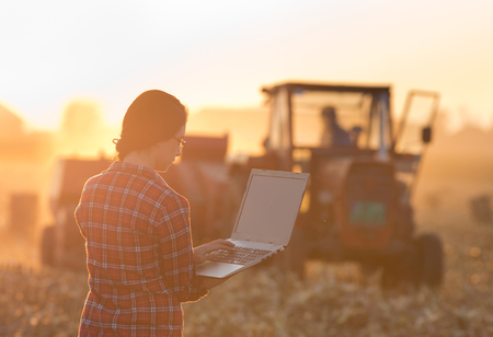 Young woman with laptop standing on field in sunset while tractor baling in background
