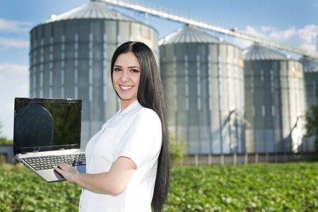 Young woman agronomist in white coat standing with laptop in front of grain silo