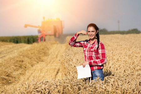 Pretty young farmer girl standing on golden wheat field. Combine harvester in backgroundの写真素材