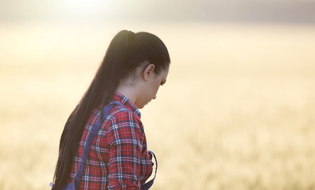 Young pretty farmer girl in plaid shirt and overalls standing in wheat field at sunset in early summer timeの写真素材