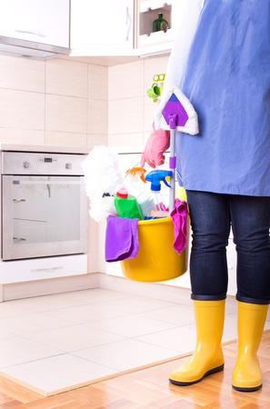 Cleaning lady with apron and gumboots holding basket full of cleaning supplies and equipment in front of kitchenの写真素材