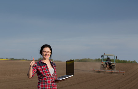 Young happy farmer girl standing on field with laptop and showing ok sign with thumb up. Tractor in backgroundの写真素材