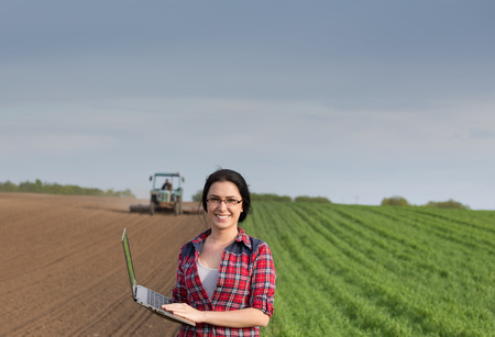 Young happy farmer girl standing on field with laptop in spring time and looking at camera. Tractor working in backgroundの写真素材