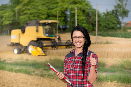Young pretty farmer girl writing notes on farmland during wheat harvest. Combine harvester in backgroundの写真素材