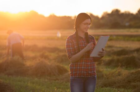 Young farmer girl writing notes on field in sunset. Old peasant working with hayfork in backgroundの写真素材