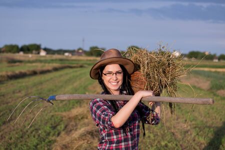 Pretty young farmer girl with hat carrying basket with hay and hayfork and smiling on the fieldの写真素材