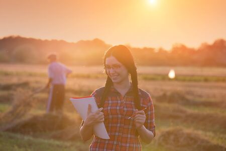 Young farmer girl writing notes on field in sunset. Old peasant working with hayfork in backgroundの写真素材