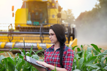 Young pretty farmer girl writing notes in corn field during wheat harvest. Combine harvester in backgroundの写真素材