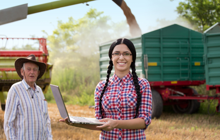 Young pretty farmer girl with laptop standing in front of combine harvester, trailer and senior man in wheat fieldの写真素材