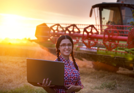 Smiling young farmer girl with laptop standing in front of combine harvester at sunset on fieldの写真素材