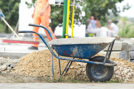 Construction wheelbarrow standing beside gravel and concrete curb stones at building site. Workers in backgroundの写真素材
