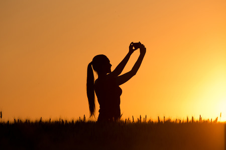 Silhouette of beautiful girl taking selfie in wheat field at sunsetの写真素材