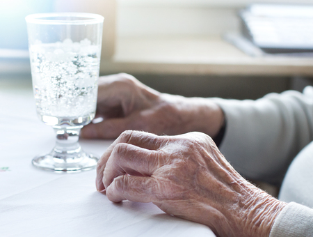 Close up of old woman's wrinkled hands on table with glass of waterの写真素材
