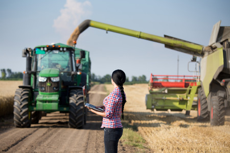 Pretty young farmer woman with laptop standing in front of tractor with trailer and combine harvester at summer field workの写真素材