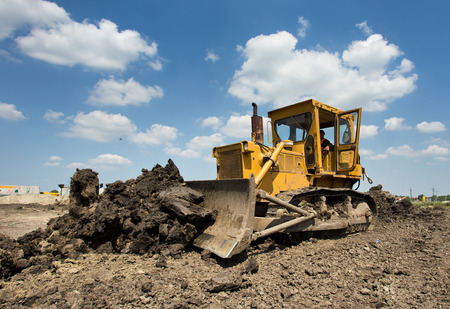 Old yellow bulldozer with caterpillar moving ground with scoop. Blue sky and white clouds in...