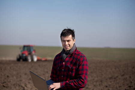 Young farmer working on laptop on field with tractor in background harrowing soil. Agricultural works in early springの写真素材