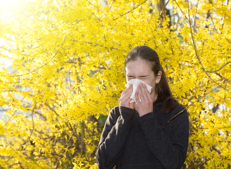 Young girl blowing nose and sneezing in tissue in front of blooming tree. Seasonal allergens affecting peopleの写真素材
