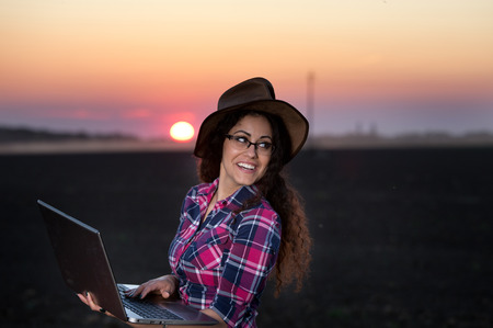 Young country woman with hat holding laptop in field at sunsetの写真素材
