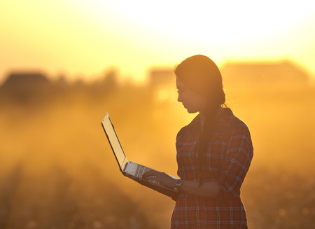 Young farmer woman with laptop standing on field at sunsetの写真素材