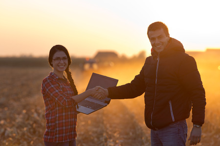 Woman engineer with laptop and landowner shaking hands on field at sunset with tractor working in background. Agribusiness conceptの写真素材