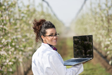 Young woman agricultural engineer in white coat with laptop standing in modern orchard with blossoming trees in spring and looking over shoulderの写真素材