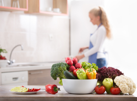 Colorful fresh vegetables in bowl on table with young blonde woman cooking in background. Healthy eating conceptの写真素材
