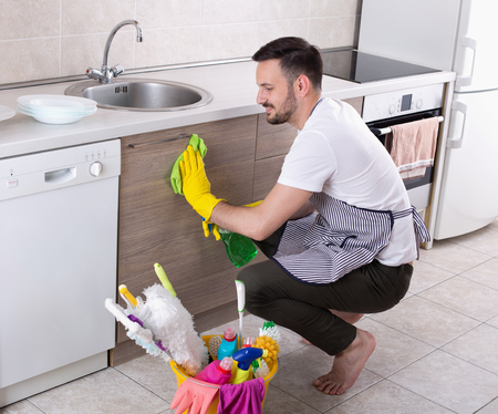 Smiling young man with apron and protective gloves wiping kitchen cabinets. Husband doing choresの写真素材