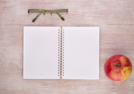 Top view of open notebook with empty pages, reading glasses and apple on wooden tableの写真素材