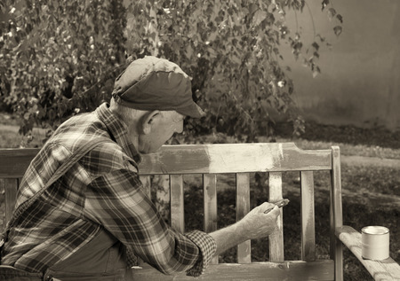 Senior man in overalls painting old bench in park after sandblasting. Repairing old furniture. Black and white imageの写真素材