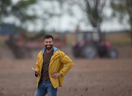 Young farmer with beard holding tablet in front of tractor in field. Seasonal agricultural works