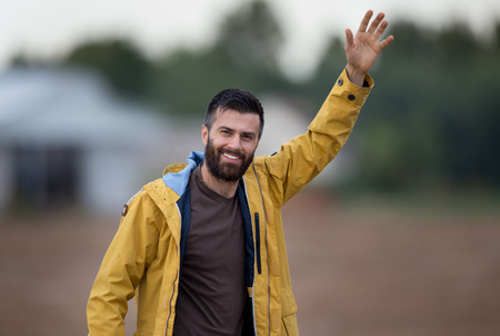 Young attractive man with beard in yellow jacket walking in field and waving handの写真素材