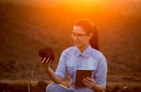 Pretty young engineer woman holding earth clod and tablet and researching soil in fieldの写真素材
