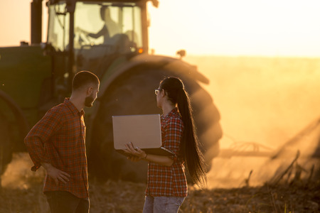 Two young farmers with laptop standing in front of tractor in field at sunsetの写真素材