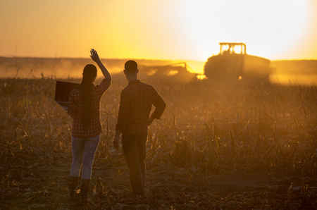 Two young farmers with laptop standing in field at sunset and looking at tractor workingの写真素材